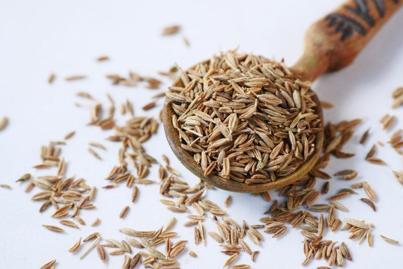 on a white surface is a wooden spoon full of cumin seeds with loose cumin seeds around it