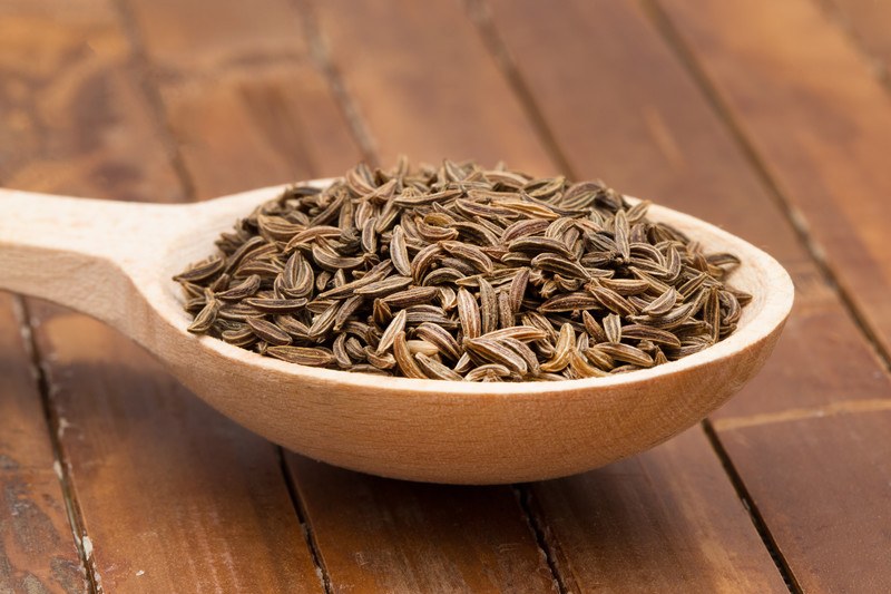 a wooden ladle full of cumin seeds resting on a wooden surface