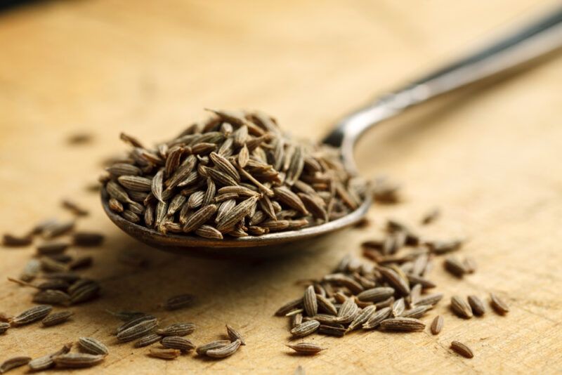 on a wooden surface is a closeup image of a silver spoon full of cumin seeds, with loose cumin seeds around it