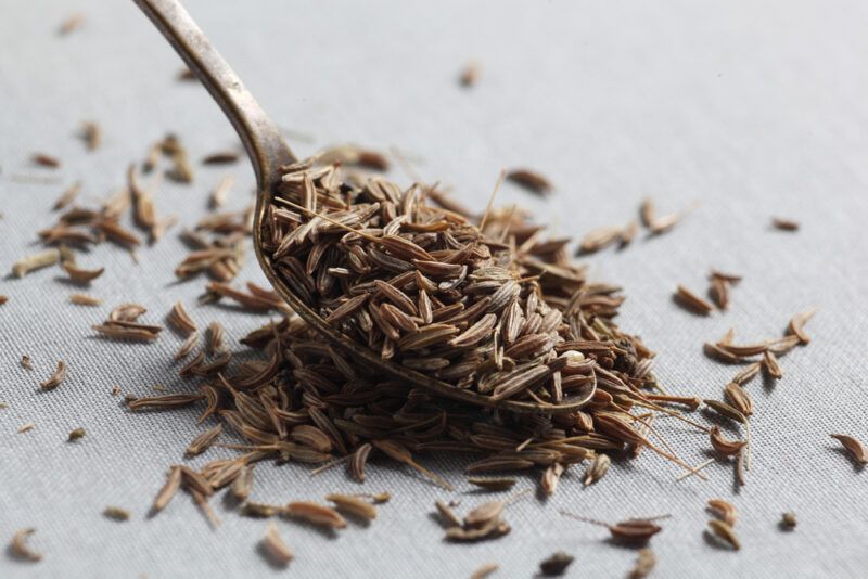 a closeup image of a spoon full of cumin seeds with loose cumin seeds around it