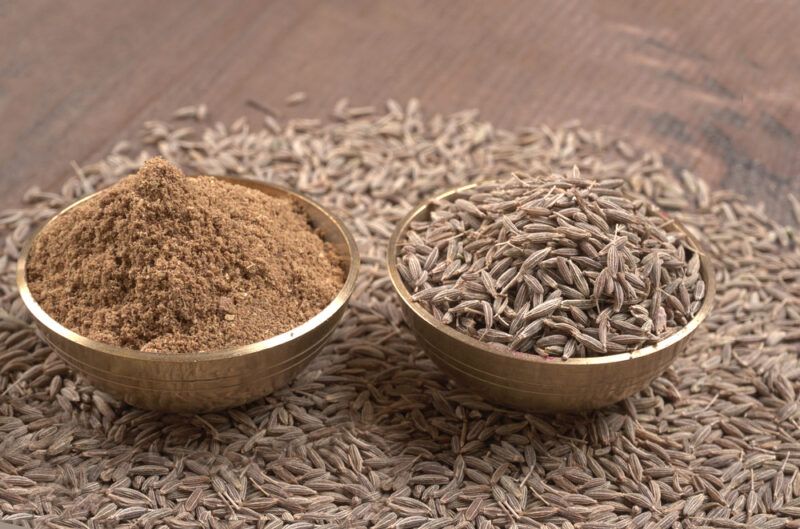 on a wooden surface is a mound of whole cumin seeds with a couple of bowls on top, one filled with whole cumin seeds, the other with ground cumin seeds