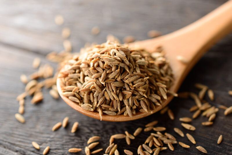 on a wooden surface is a closeup image of a wooden spoon with cumin seeds with loose cumin seeds around it