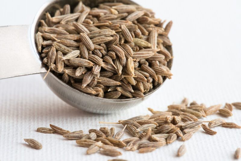 on a white table napkin is a closeup image of a metal measuring spoon full of cumin seeds, around it are loose cumin seeds