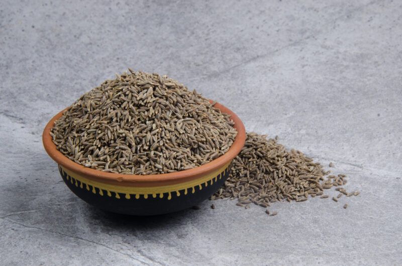 on a concrete surface is a black and brown ceramic bowl full of cumin seeds, at the back is a small mound of cumin seeds