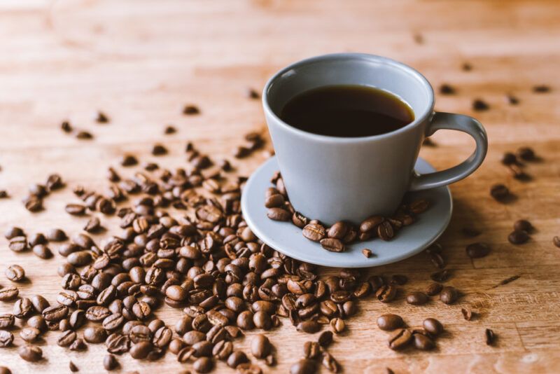 on a wooden surface is a bluish cup of black coffee with loosed whole coffee beans around it