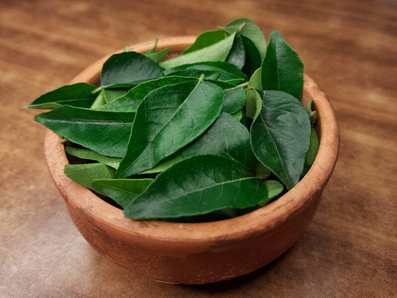 on a dark wooden surface is a clay bowl full of fresh curry leaves