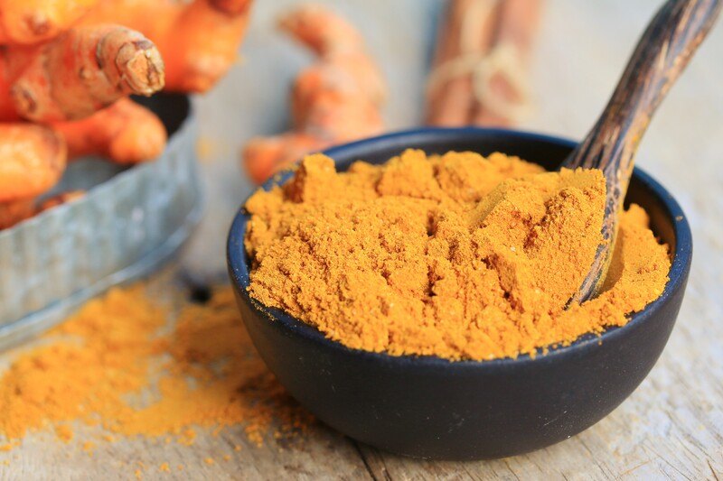 on a wooden surface is a black bowl full of curry powder with a wooden spoon, behind it are fresh turmeric and loose curry powder