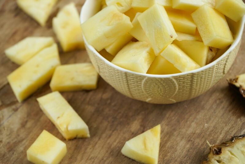 closeup image of a bowl full of cubed pineapple bits, resting on top of a wooden board, around it are loose cube pineapple bits as well