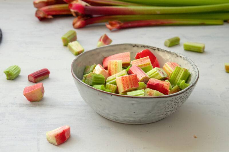 on a beige colored surface is a bowl full of cut rhubarb, around it are loose cut rhubarb and a long stalks at the back