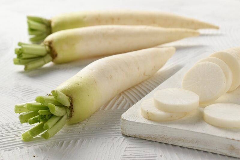 on a white surface are three whole daikon with a white wooden chopping board with sliced daikon on it