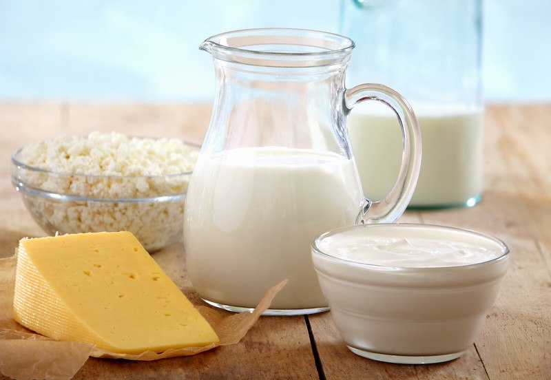 This photo shows a pitcher of milk, a pitcher of cream, a dish of yogurt, a dish of cheese curds, and a wedge of orange cheese on a wooden table.