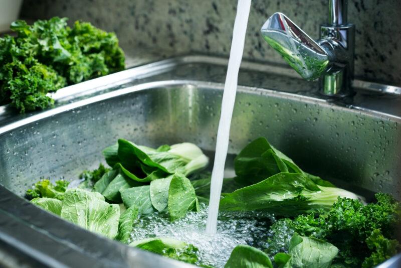 different dark leafy green vegetables in the sink with the water faucet running