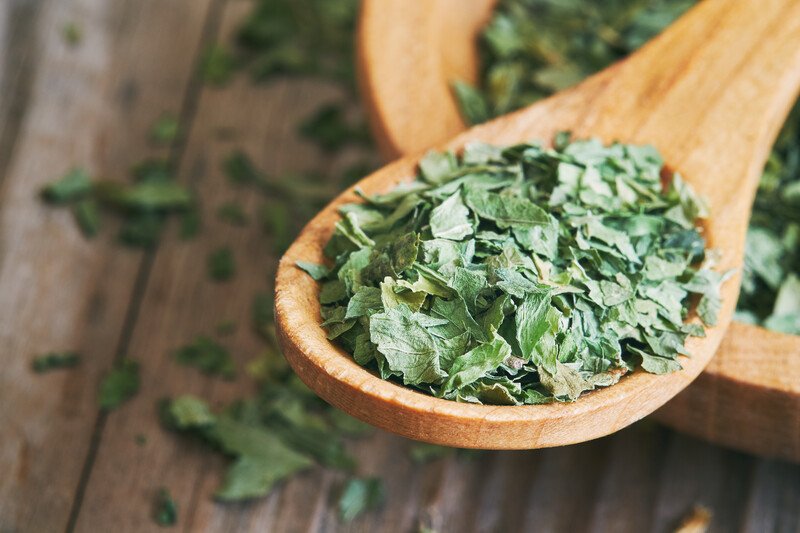 on a wooden surface is a closeup image of a wooden spoon full of dehydrated celery  resting on a wooden bowl full of dehydrated celery, with loose dehydrated celery around it