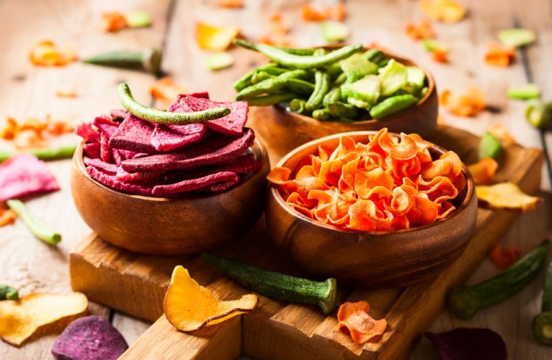 Three bowls f dried vegetables in orange, green, and maroon rest on a cutting board on a wooden surface near loose dried vegetables.