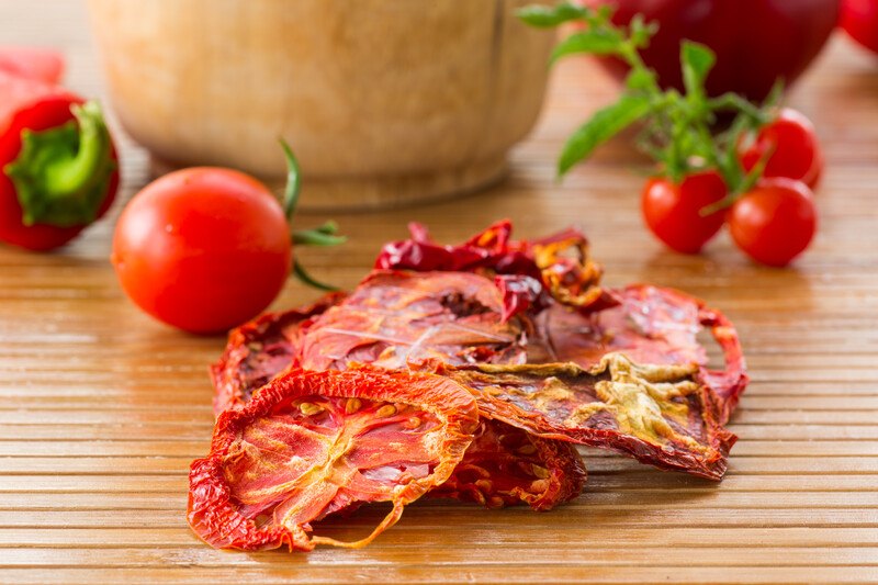 on a wooden surface is a closeup image of dehydrated tomatoes with fresh tomatoes and cherry tomatoes at the back