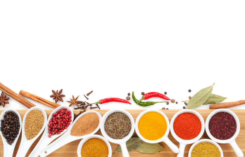 peppers, cumin, rosemary, and other spices on white spoons placed on top of a wodden board.