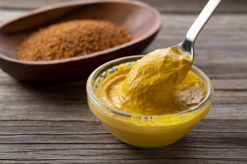 on a dark wooden surface is a clear bowl of Dijon mustard, at the back is a brown dish with ground brown spice