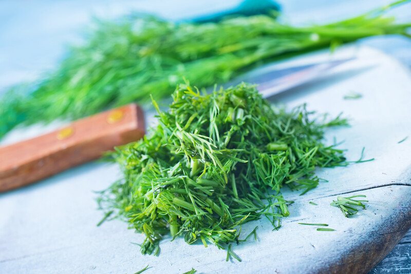 a white wooden chopping board with chopped dill, knife and fresh dill at the back