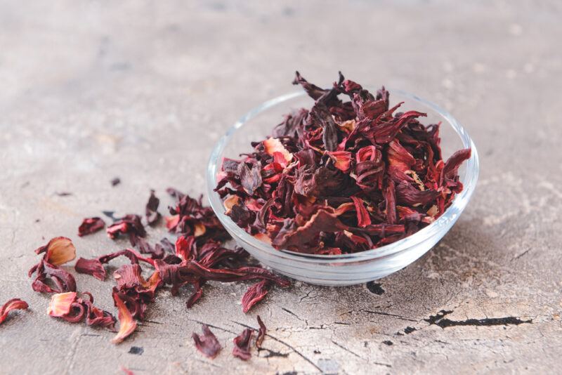 on a concrete surface is a clear glass bowl of dried hibiscus with loose dried hibiscus beside beside it