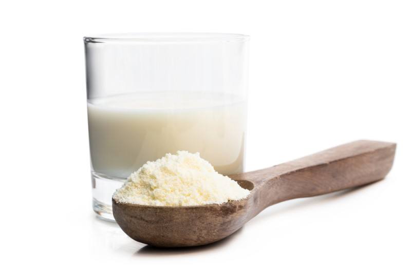 A wooden spoon with powdered milk in it rests in front of a clear glass with milk in it, against a white background.