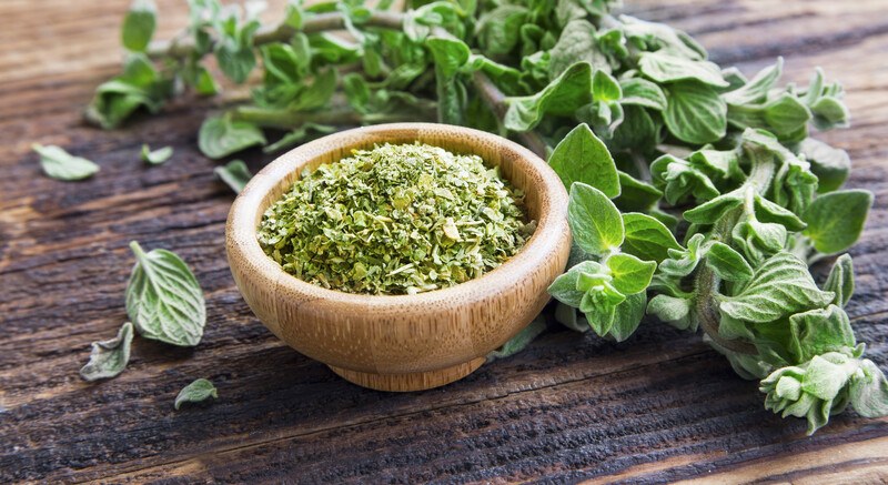 on a dark wooden surface is a wooden bowl full of dried oregano with fresh oregano leaves at the back