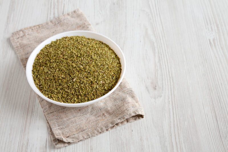 on a very light wooden surface is a white bowl full of dried oregano resting on a light brown table napkin