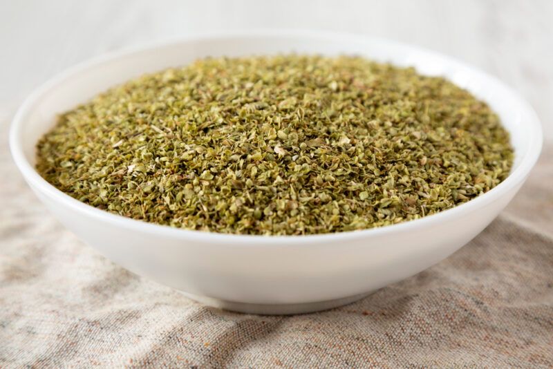 closeup image of a white ceramic bowl full of dried oregano resting on top of a tan colored table napkin