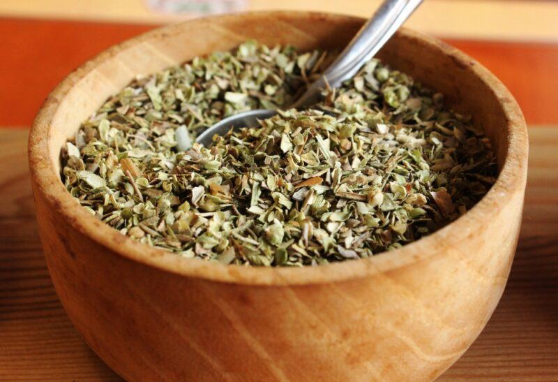 closeup shot of a wooden bowl full of dried oregano with a silver spoon shoved in it