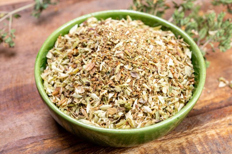 on a wooden surface is a green ceramic bowl full of dried oregano, at the back are dried sprigs of oregano