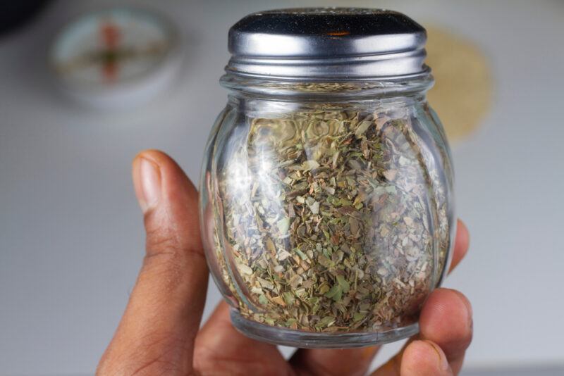 a closeup image of a hand holding a small spice jar with dried oregano