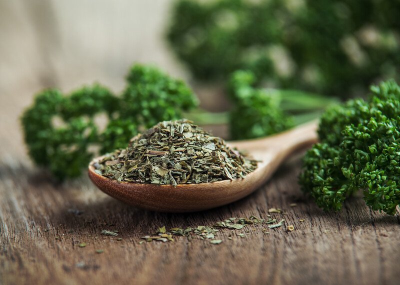 on a wooden surface is a wooden ladle full of dried parsley, around it are fresh parsley