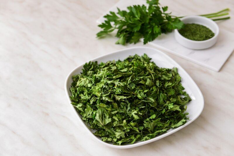 on a white marbled surface is a white square dish with dried parsley, at the back is a small round dish of dried parsley with a sprig of fresh parsley resting on a rectangle marble board