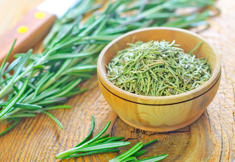 on a wooden surface is a wooden bowl full of dried rosemary with fresh rosemary herbs around it