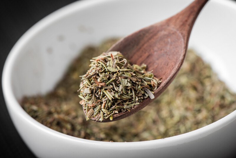 a closeup image of a white bowl with dried thyme at the back, focus is on the wooden spoon with dried thyme as well