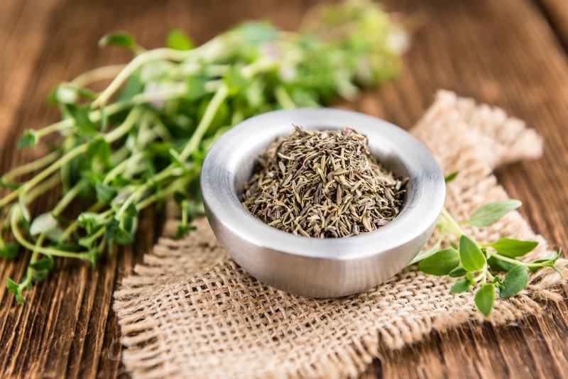 on a wooden surface is a metal bowl full of dried thyme resting on top of a piece of burlap with fresh thyme at the back