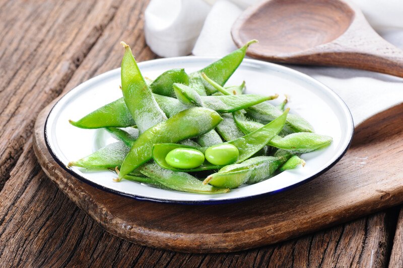 on a rustic wooden surface is a wooden board with a plate of green edamame with a wooden ladle at the back