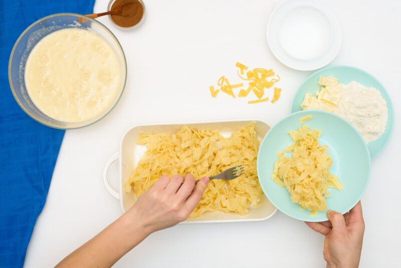 overhead shot of ingredients including lokshen noodles, cream, sugar, and more, and a couple of hands making kugel