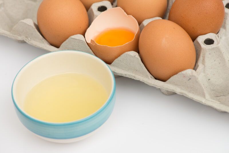 on a white surface is a white and blue bowl with egg white, at the back is a carton of brown eggs with one open in half with a yolk in it