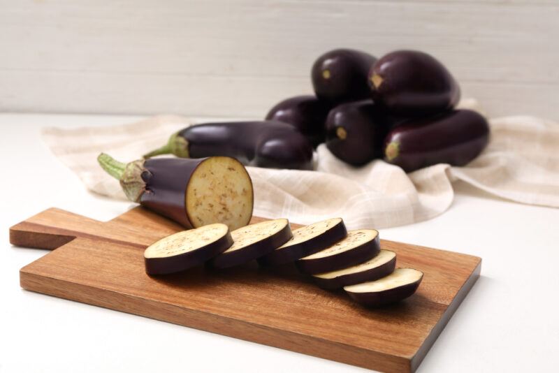 on a white surface is a wooden chopping board with sliced eggplant, at the back are whole eggplants resting on top of a white kitchen towel