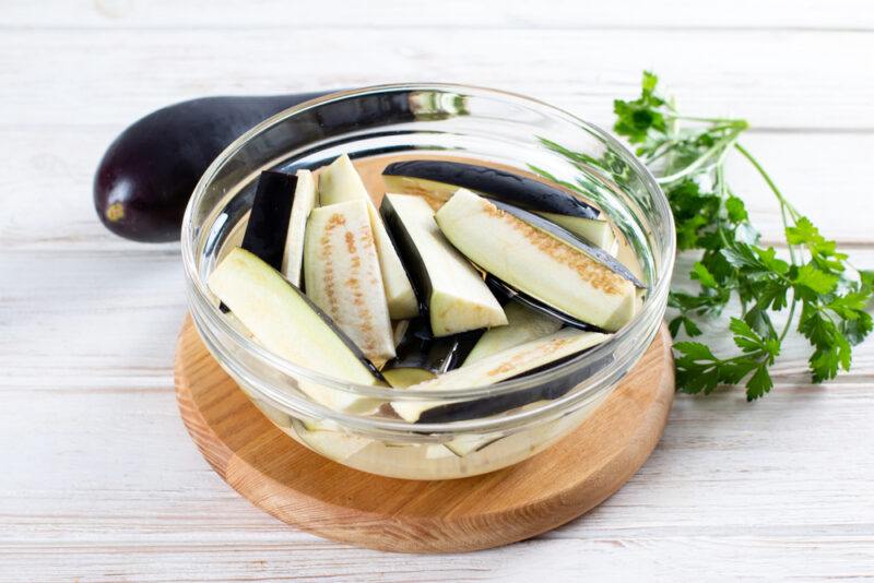 on a white wooden surface is a round brown wooden board with a clear glass bowl of sliced eggplants, at the back is a couple of fresh eggplant and a sprig of cilantro