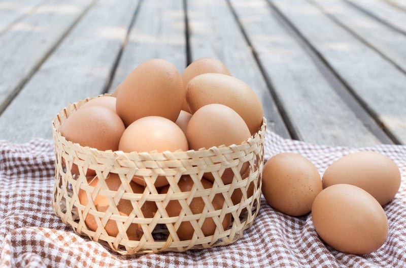 on an aged wooden surface is a basket full of brown eggs and 3 eggs beside it, resting on a checkered table napkin