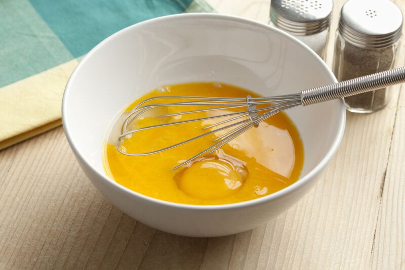 partially beaten eggs in a white bowl with metal whisk, resting on a wooden surface with salt and pepper shaker behind it