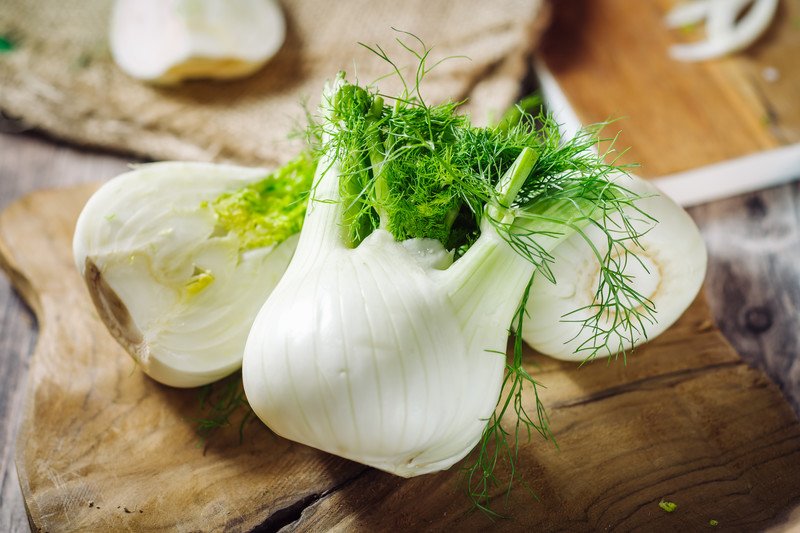 closeup image of a couple of whole fennel bulb and half bulb, all fennel are laid out on a wooden chopping board, with cut fennel at the back on a burlap sack