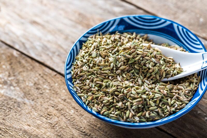 on a wooden surface is a blue glass bowl with prints full of fennel seeds with a ceramic spoon shoved in it