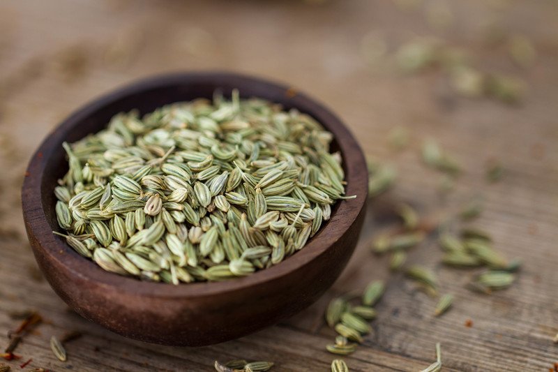 closeup image of a dark wooden bowl full of fennel seeds resting on a wooden surface with loose fennel seeds around it