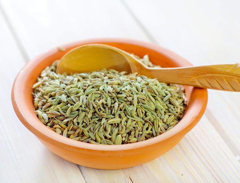 on a white wooden surface is a brown ceramic bowl full of fennel seeds with wooden spoon