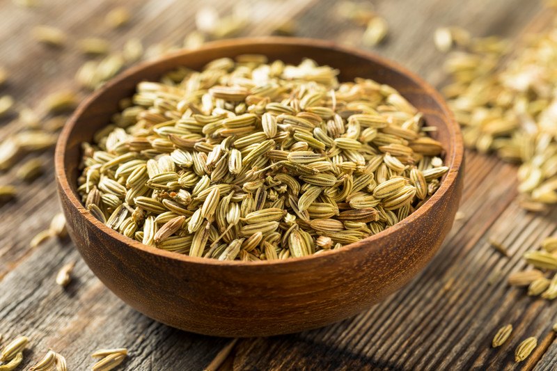 on a wooden surface is a brown wooden bowl full of fennel seeds with loose fennel seeds around it