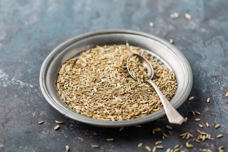 on a gray surface is a metal dish full of fennel seeds with a silver spoon