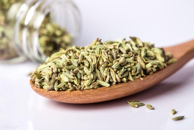 on a white surface is a closeup image of a wooden spoon full of fennel seeds, behind it is a small jar topped over with fennel seeds pouring out from it