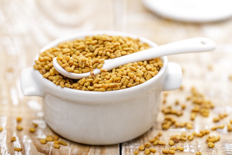 on a wooden surface is a white ceramic bowl with fenugreek seeds with ceramic spoon on top, and loose fenugreek seeds around it